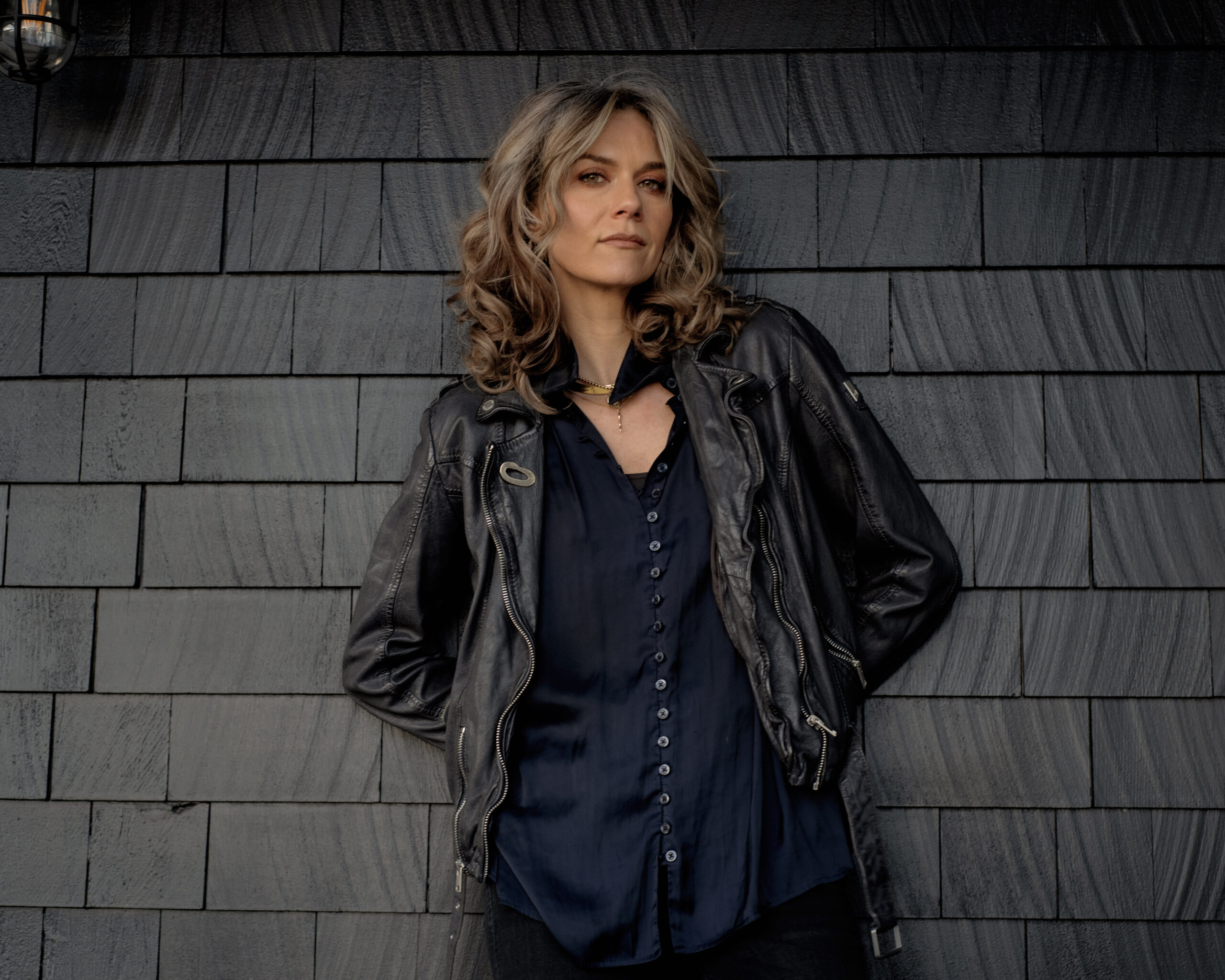 A woman with wavy, shoulder-length hair stands against a dark shingle wall, wearing a black leather jacket over a navy blue button-up shirt, looking confidently at the camera.