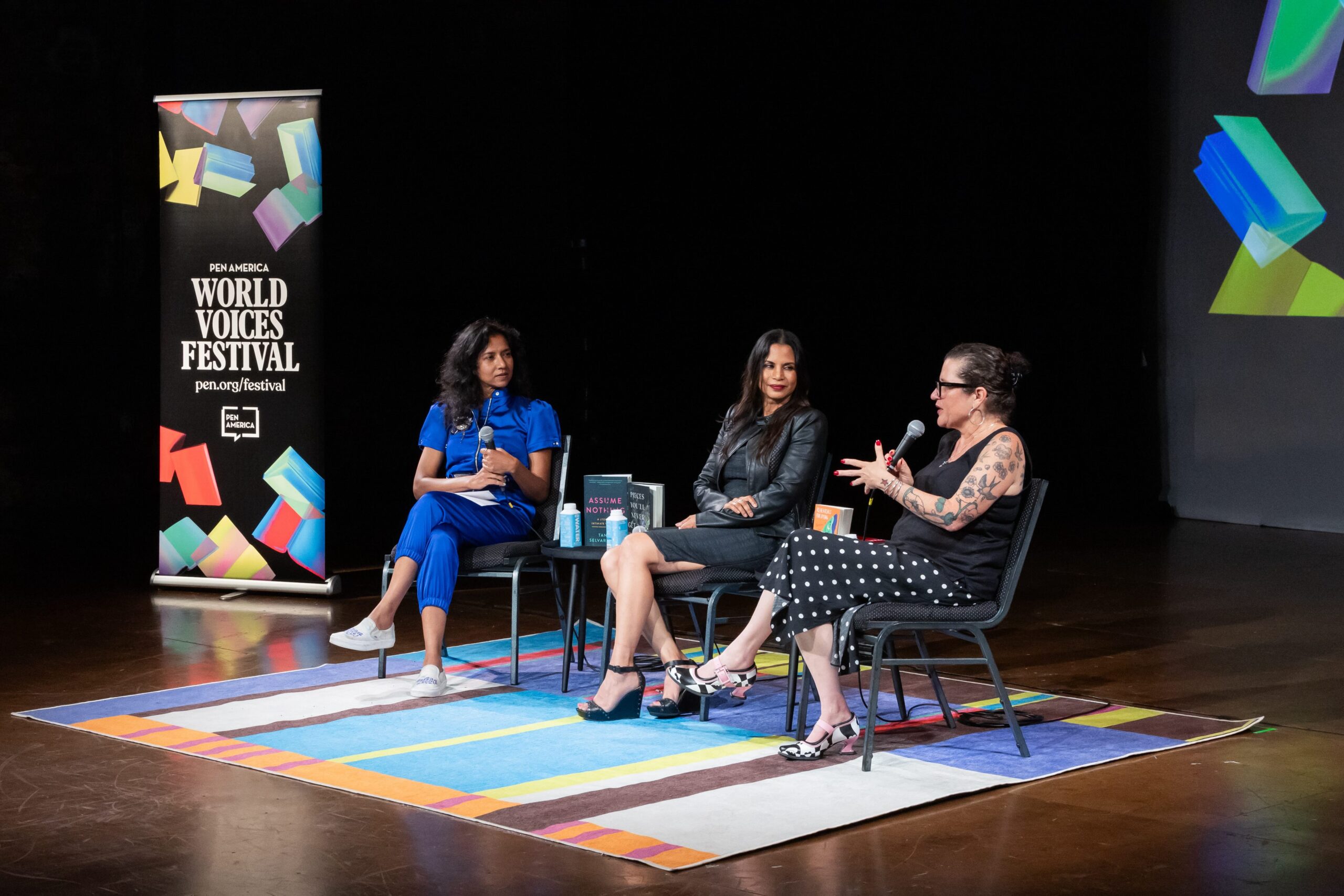 Three women sit on stage at the PEN America World Voices Festival, engaged in a panel discussion. Two listen while one, wearing glasses and patterned dress, gestures as she speaks. Colorful graphics are displayed in the background.