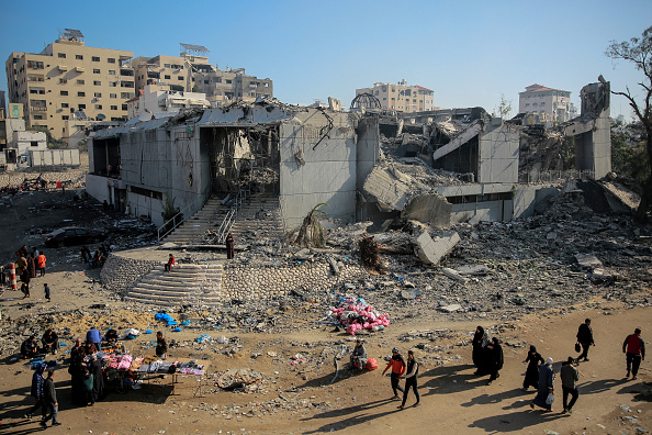 People walk near a heavily damaged building surrounded by rubble in an urban area. Makeshift stalls with goods are visible in the foreground, and several buildings stand in the background under a clear sky.