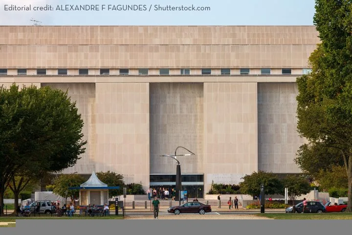 A large, modern beige stone building with tall vertical columns and small upper windows, surrounded by trees and people walking in front. Several cars are parked nearby.