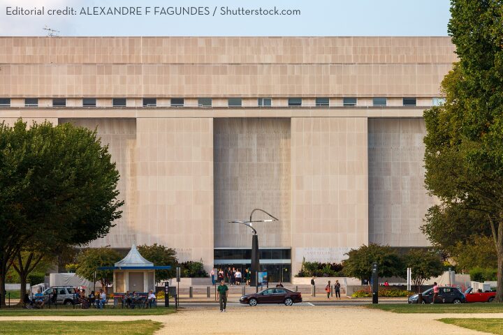 A large, modern beige stone building with tall vertical columns and small upper windows, surrounded by trees and people walking in front. Several cars are parked nearby.