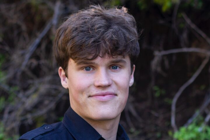 A young man with wavy brown hair and blue eyes smiles slightly while standing outdoors in front of a blurred background of branches and greenery. He is wearing a dark collared shirt.