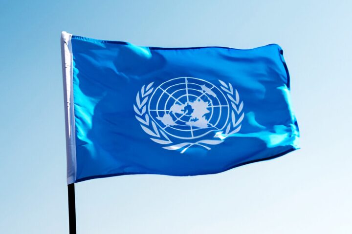 A blue flag with the white emblem of the United Nations, featuring a world map surrounded by olive branches, waves against a clear blue sky.