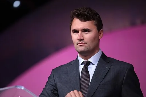 A man in a dark suit and tie stands at a podium against a pink and purple background, looking forward with a neutral expression.