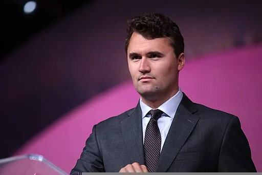 A man in a dark suit and tie stands in front of a purple and pink background, looking forward with a neutral expression.