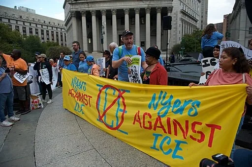 A group of people at a protest hold a large yellow banner reading NYers Against ICE with a crossed-out ICE symbol, while others hold signs in front of a government building.