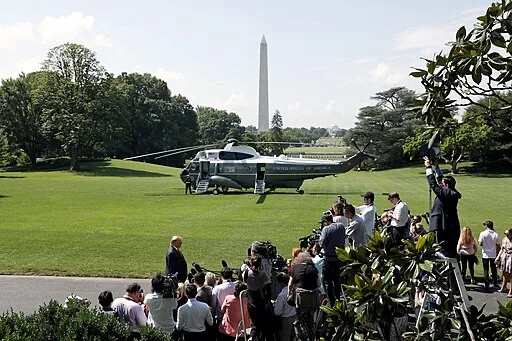 A helicopter labeled “United States of America” is parked on the White House lawn as photographers and people gather nearby, with the Washington Monument visible in the background.