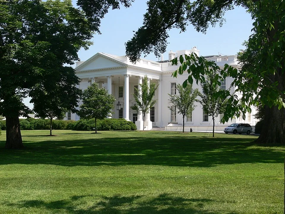 The White House is seen through leafy trees, with its white columns and portico visible. A car is parked nearby on the lush green lawn under a clear blue sky.