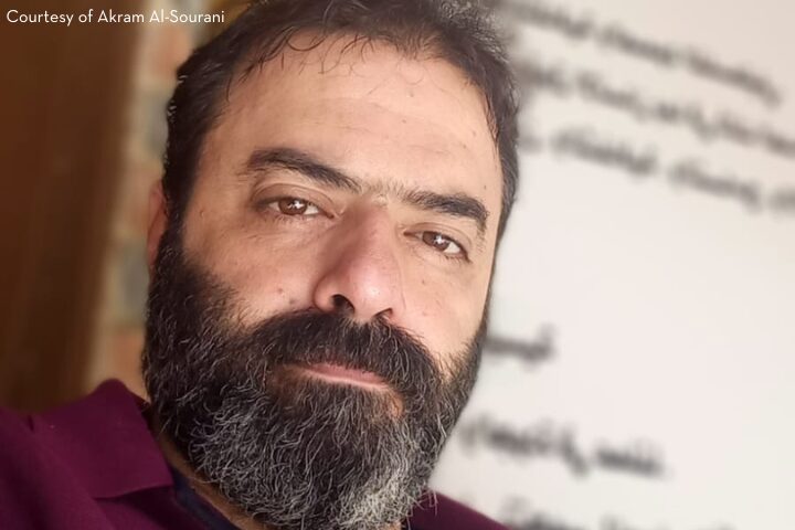 A man with a beard and mustache looks at the camera, standing indoors in front of a white wall with partially blurred Arabic writing. Courtesy of Akram Al-Sourani appears in the top left corner.