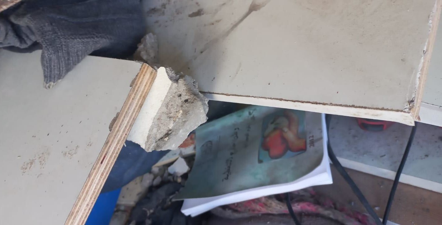 A broken wooden shelf with rough edges lies on a cluttered surface. Below it, a book with an illustrated cover and Arabic text is partially visible among scattered debris and other items.