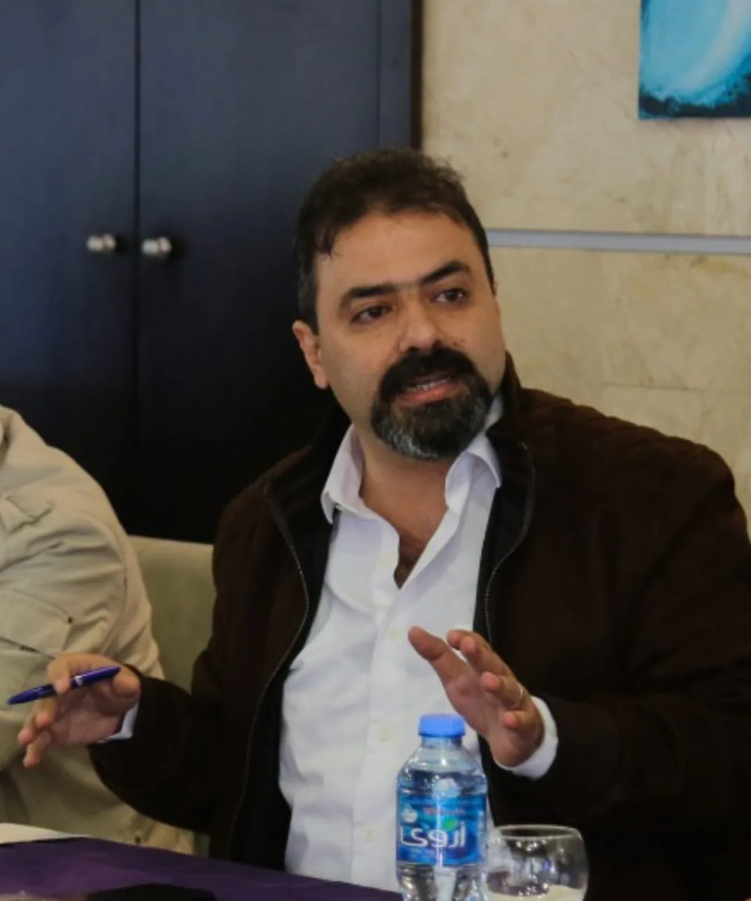 A man with dark hair and a beard, wearing a white shirt and brown jacket, gestures with one hand while holding a pen. He is seated at a table with a water bottle and glass in front of him.