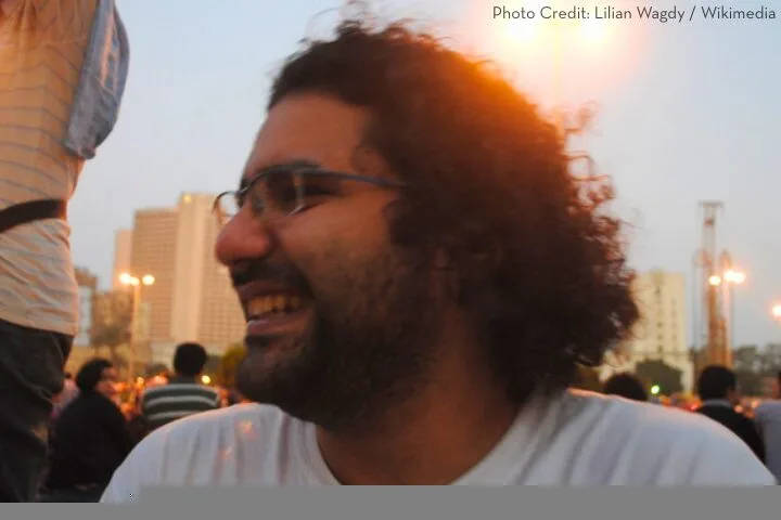 A man with curly hair and glasses smiles while sitting outdoors in a city at dusk, with blurred buildings and people in the background.