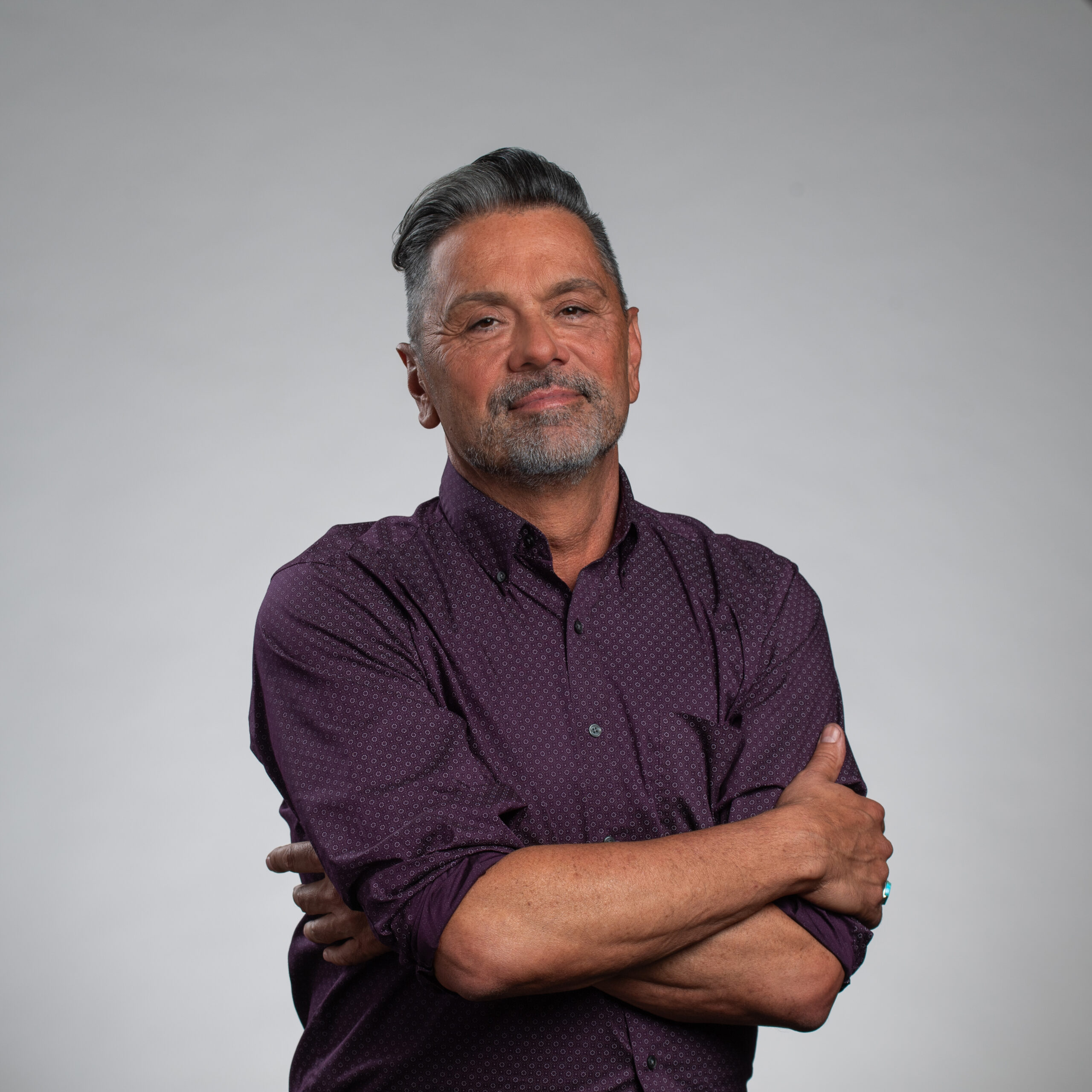 A middle-aged man with short gray hair and a trimmed beard stands against a plain gray background, wearing a purple button-up shirt and crossing his arms, smiling slightly.