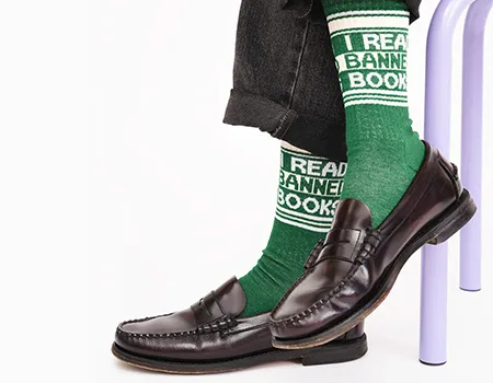 A person wearing green socks that say I READ BANNED BOOKS, paired with black loafers and dark pants, sits with legs crossed on a light purple chair against a white background—shop PEN America merchandise to show your support.