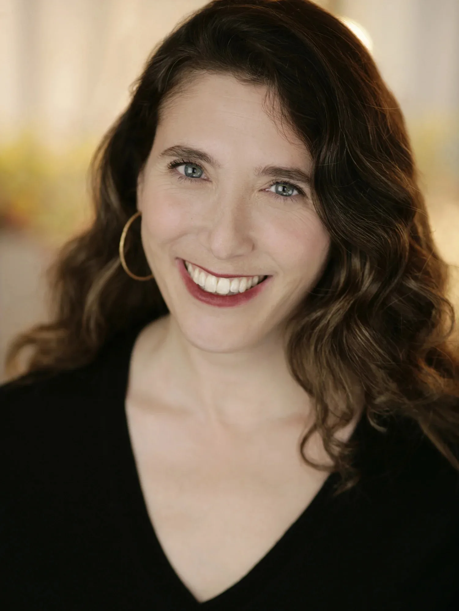 A woman with wavy brown hair and hoop earrings smiles warmly at the camera, wearing a black top against a softly blurred background.