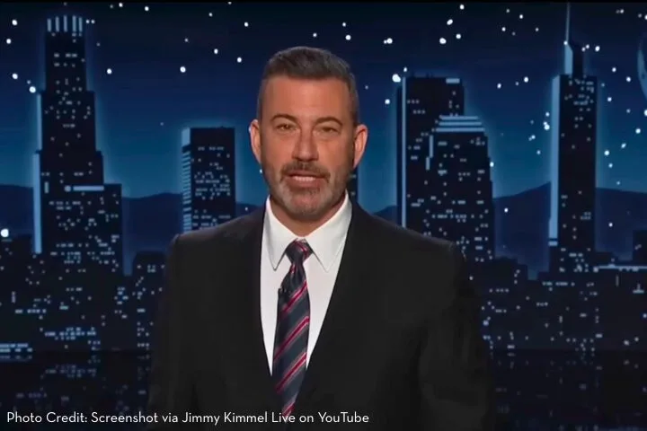 A man in a dark suit and striped tie stands on a stage with a city skyline backdrop and starry night sky. Text at the bottom credits Jimmy Kimmel Live on YouTube.