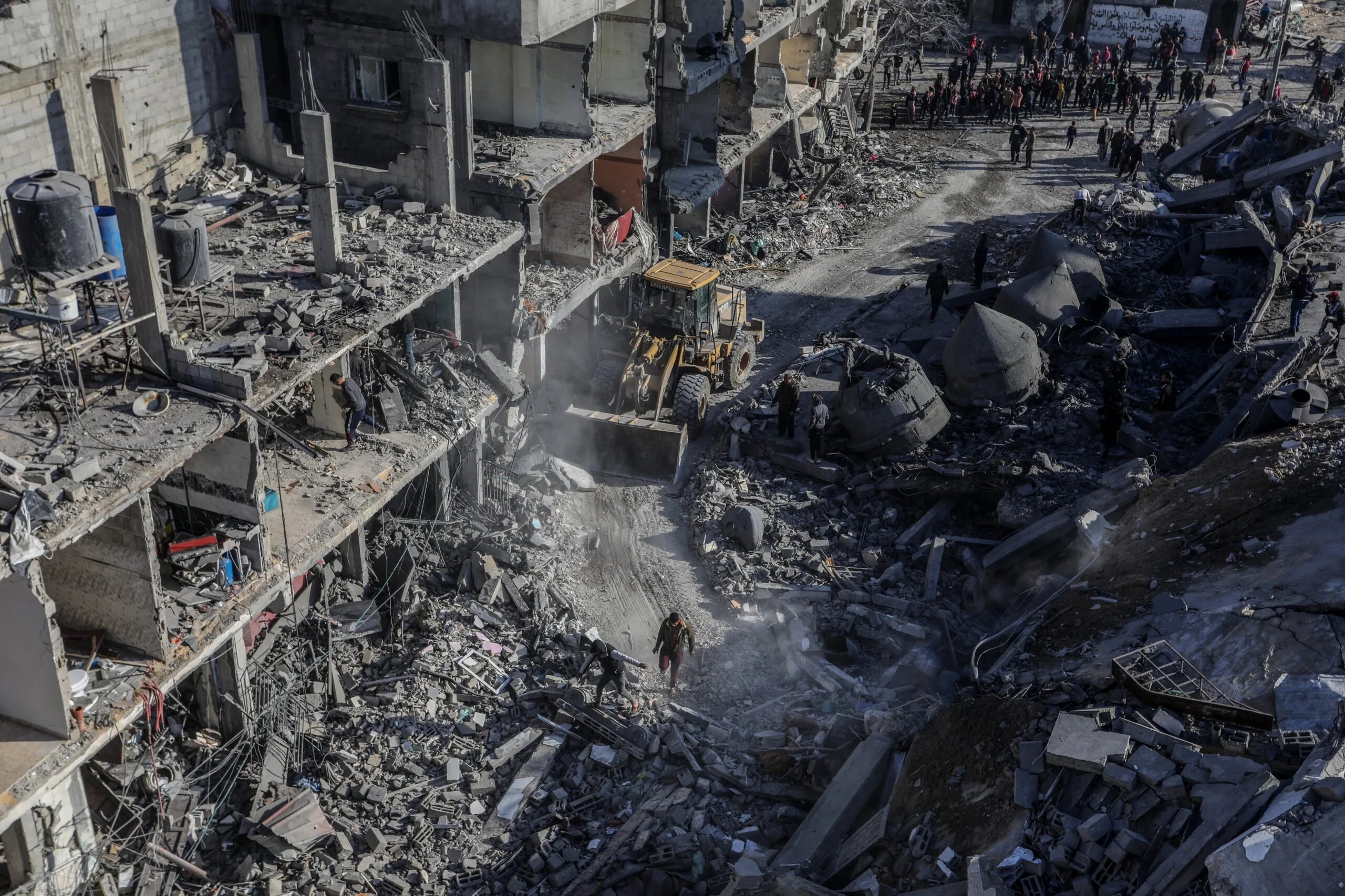 A bulldozer clears rubble from heavily damaged buildings and debris-strewn streets, while people survey the destruction and gather in the background.