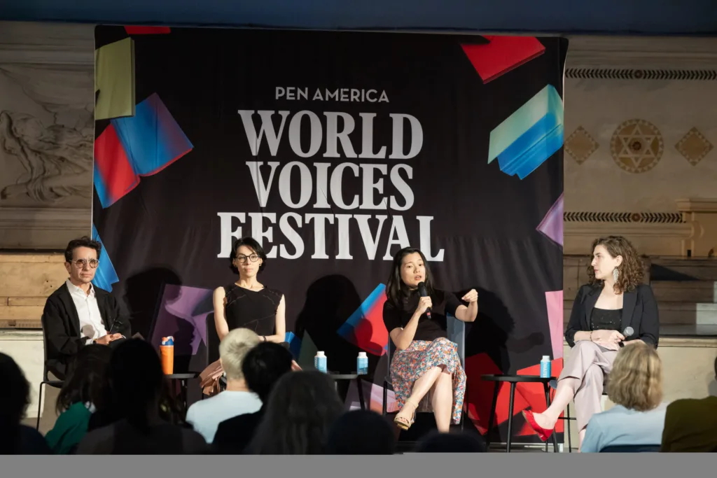 Four panelists sit on stage in front of a PEN America World Voices Festival banner, engaging with an audience. One woman speaks into a microphone while the others listen attentively.