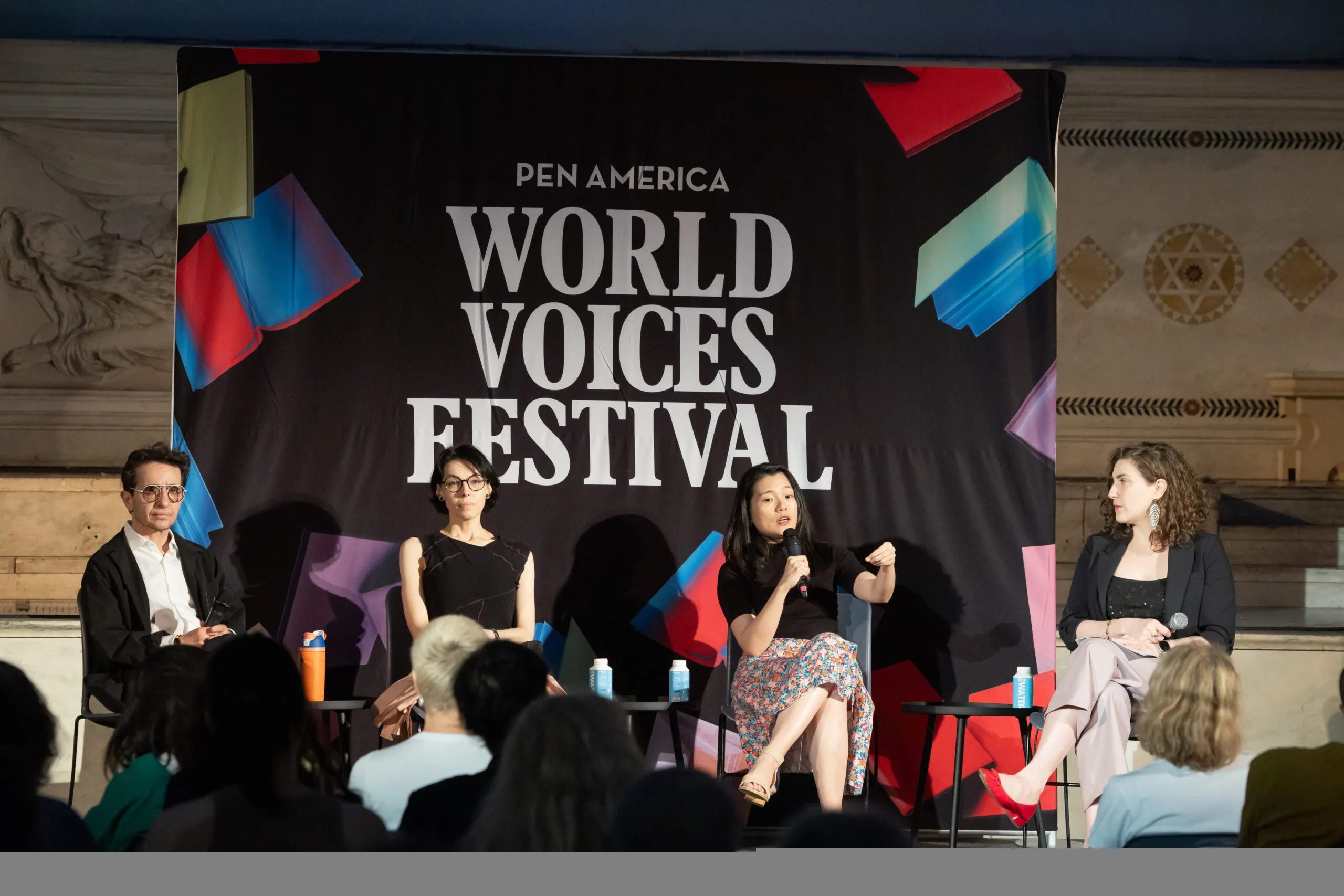 Four panelists sit on stage in front of a PEN America World Voices Festival banner, engaging with an audience. One woman speaks into a microphone while the others listen attentively.