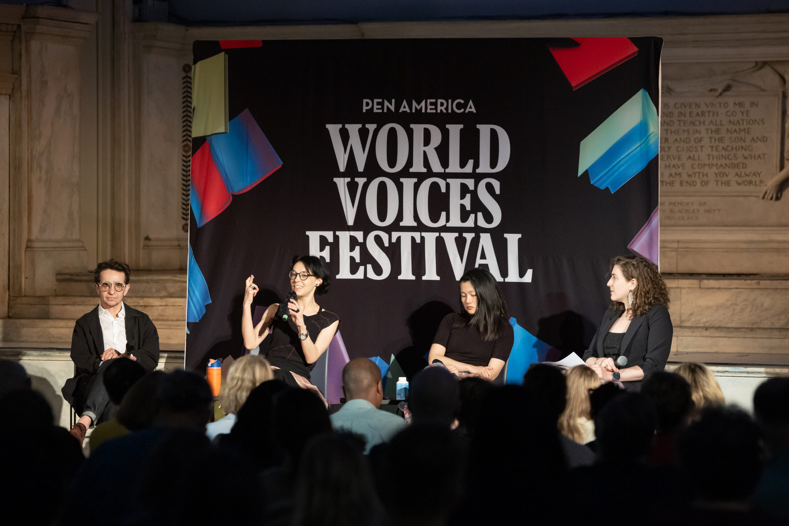 Four panelists sit on stage in front of a PEN America World Voices Festival banner, engaging with an audience, with one person signing in American Sign Language.