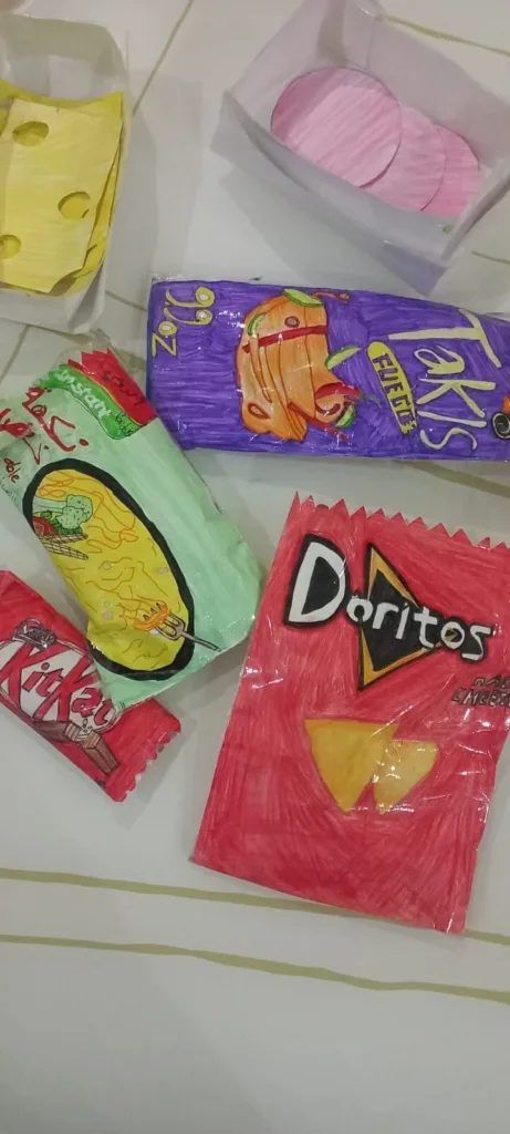 Hand-drawn paper replicas of snack packages, including Doritos, KitKat, Takis, a green chip bag, and a yellow cheese wedge, are displayed on a white surface with lines.