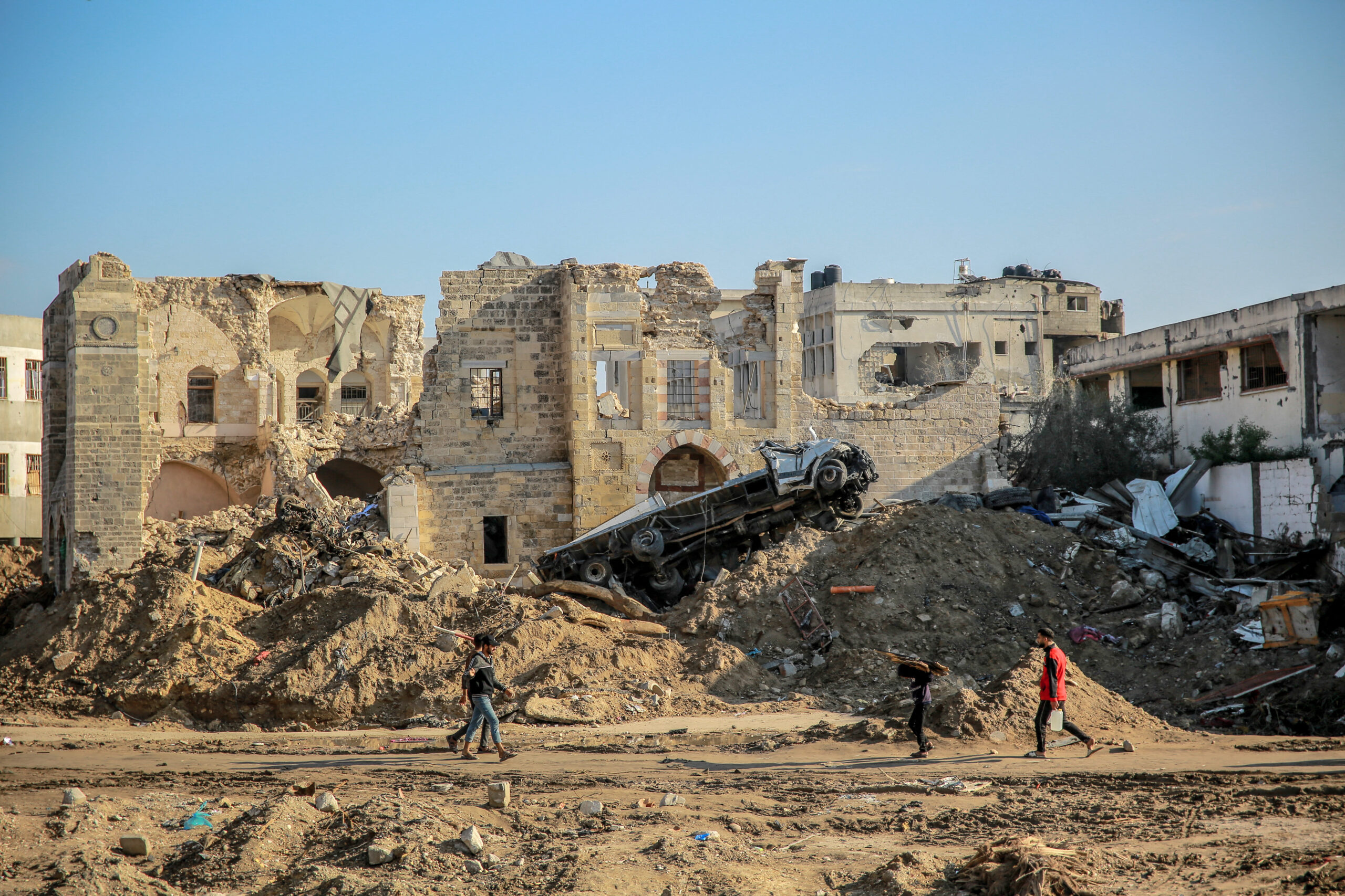 Four children walk past rubble and debris, with a destroyed building and an overturned vehicle in the background under a clear blue sky.