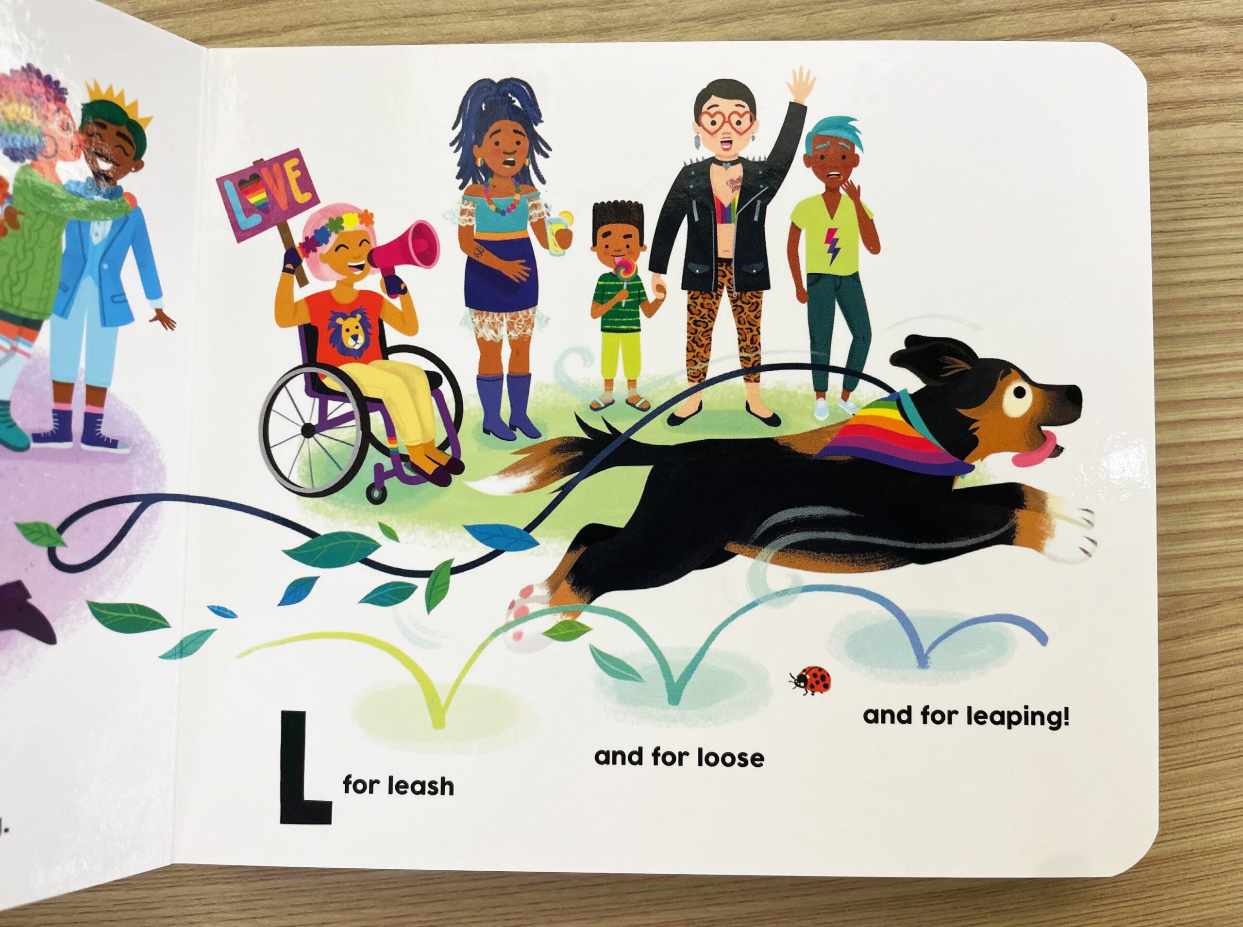 A joyful dog wearing a rainbow bandana leaps across grass, pulling a leash. Diverse people, including a person in a wheelchair holding a “LOVE” sign, cheer in the background. Text reads: L for leash and for loose and for leaping!.