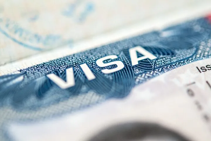 Close-up of a travel visa document, showing the word VISA in bold white letters on a blue patterned background. The details of the visa are slightly blurred in the foreground and background.