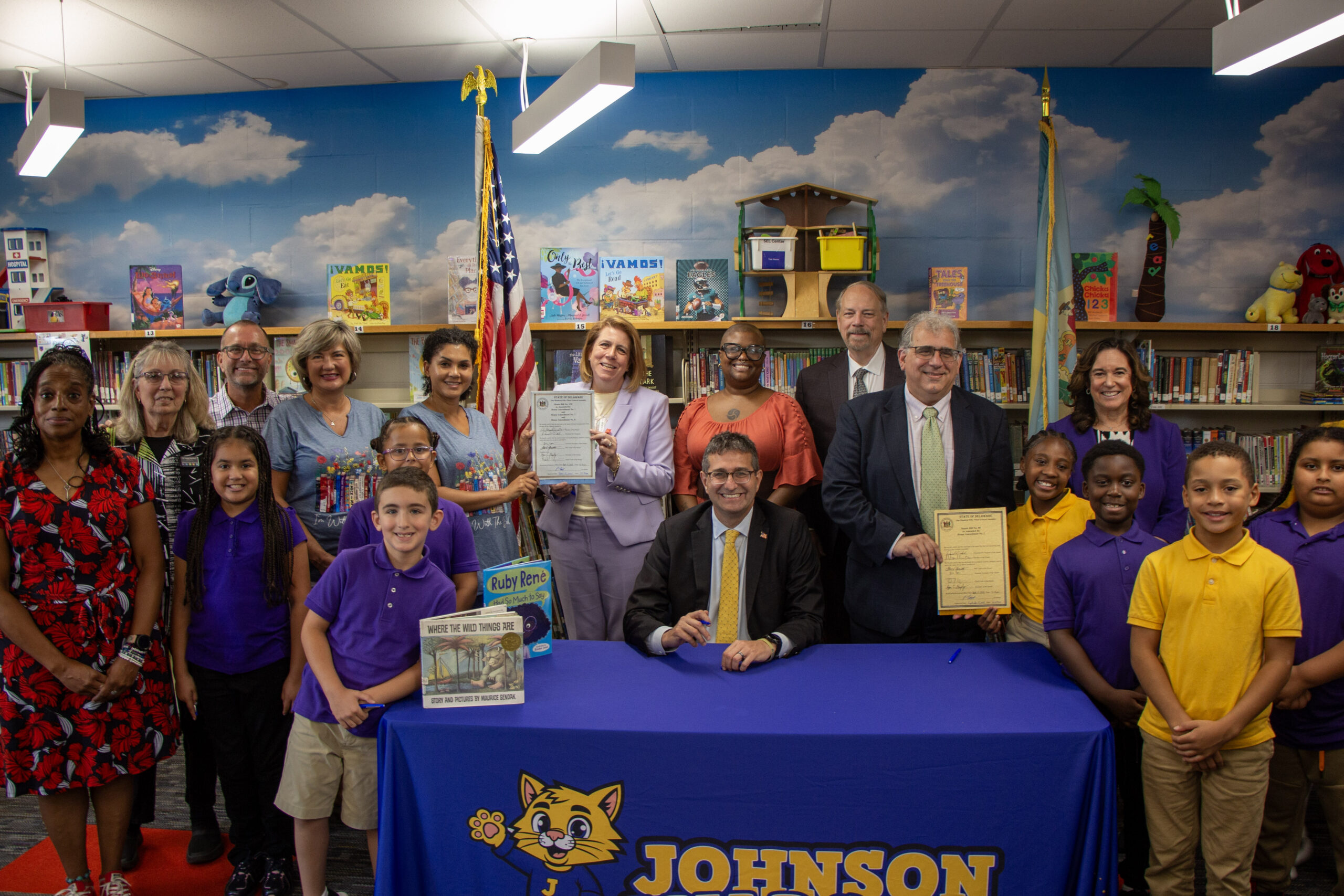 A group of adults and children pose in a colorful library with books and an American flag. Several adults hold official documents, and two men sit at a table with a Johnson sign. Everyone is smiling.