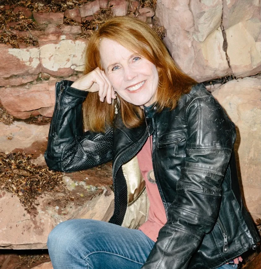 A woman with red hair wearing a black leather jacket and blue jeans sits on stone steps, smiling and resting her head on her hand, with a rocky background behind her.