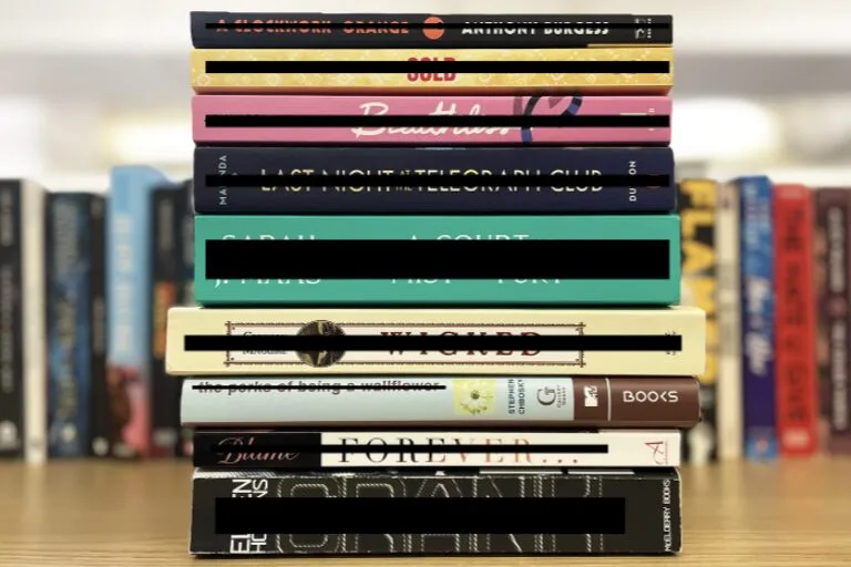 A stack of twelve books of various colors and sizes, arranged vertically on a wooden surface, with blurred bookshelves in the background. The book titles are partially obscured by black bars, suggesting they belong to a banned book list.