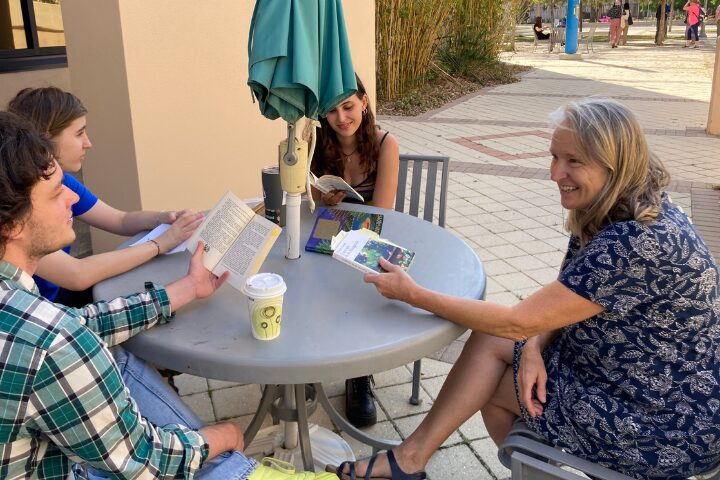 Four people sit around an outdoor table, reading and talking together. One woman holds a book and smiles, while others read and discuss. An umbrella shades the table, and a coffee cup sits nearby.