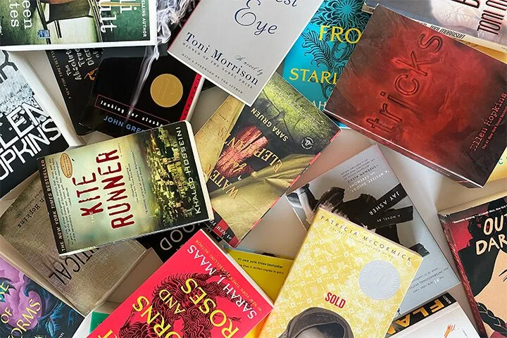 A scattered collection of various paperback and hardcover books with colorful covers, including titles like “The Kite Runner,” “Tricks,” “The Bluest Eye,” and others, laid out on a flat surface.