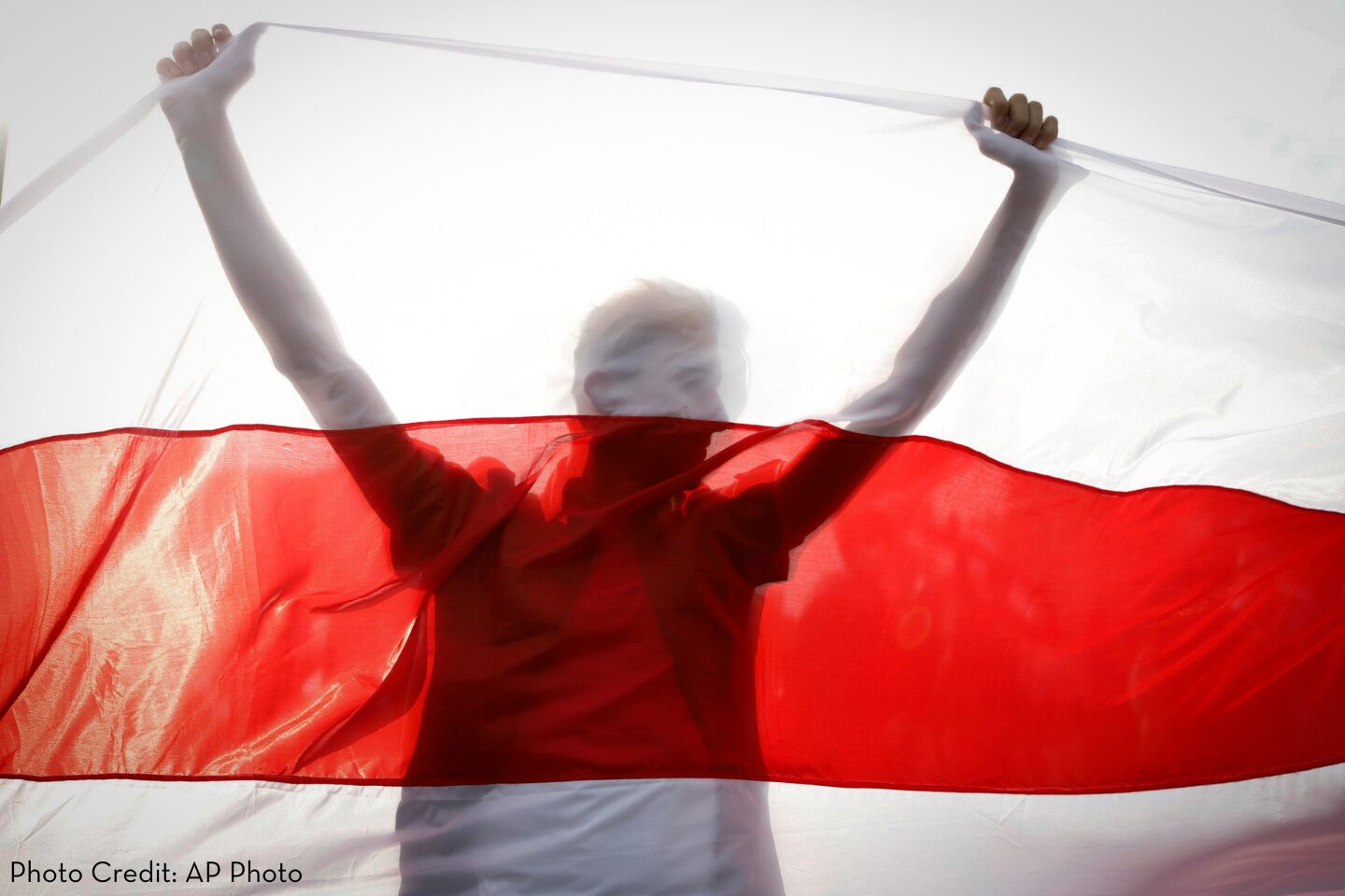 A person holds up a large white and red flag, with their silhouette visible through the fabric. The lighting is bright, creating a translucent effect. Photo Credit: AP Photo.