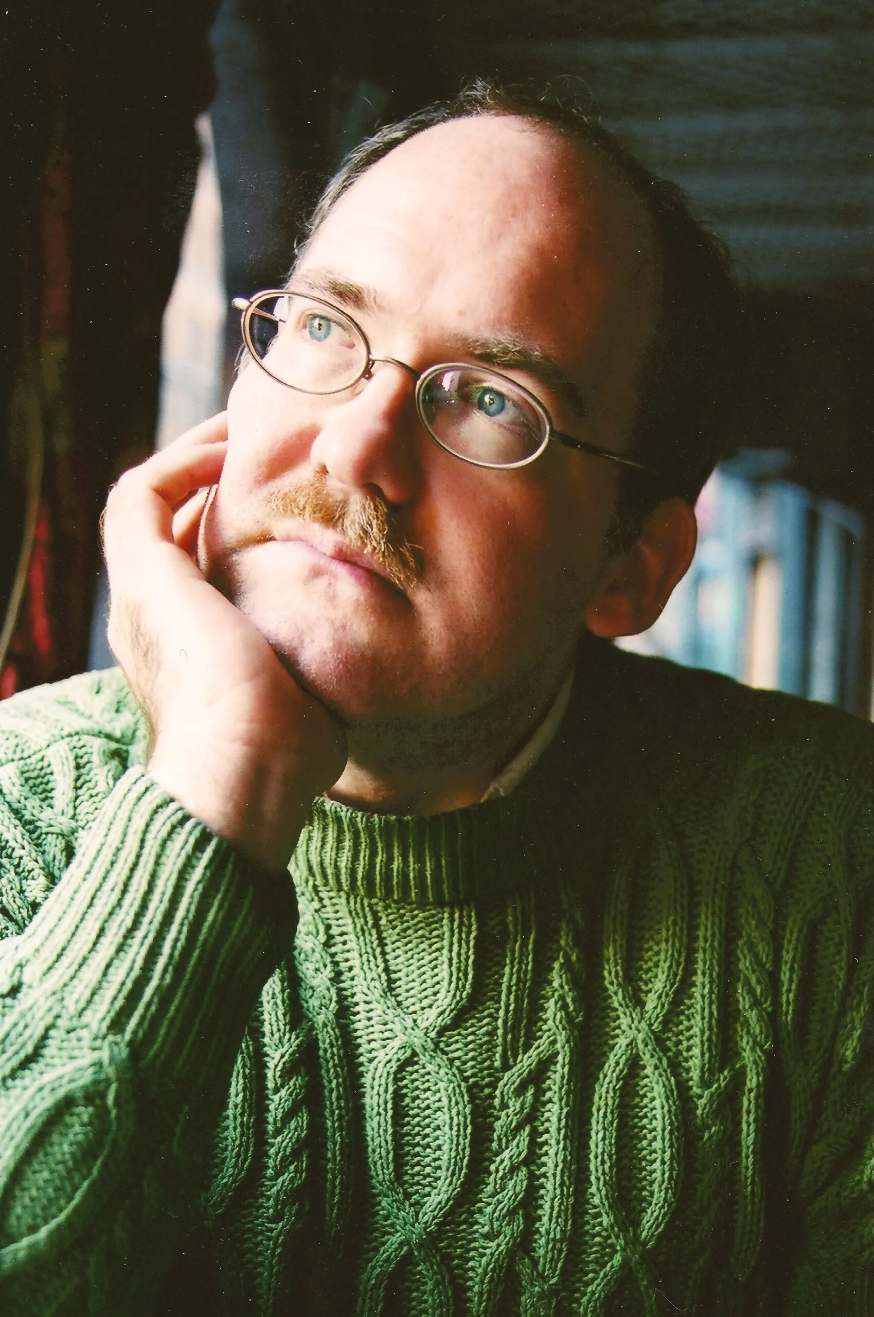 A man wearing glasses and a green cable-knit sweater rests his chin on his hand and gazes thoughtfully upwards, with soft natural light illuminating his face.