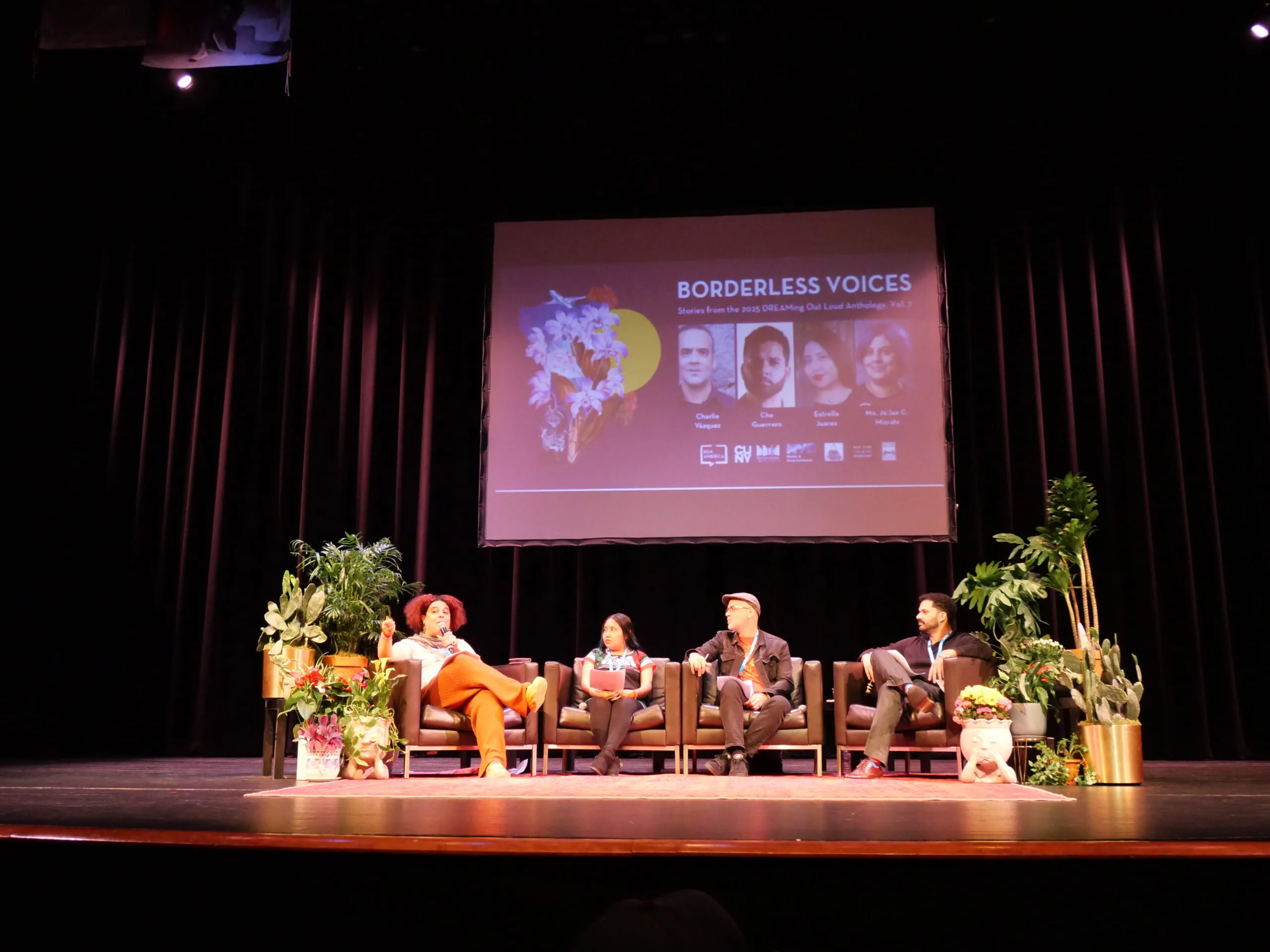 Four people sit on a stage panel discussion with plants around them. Behind them is a large screen displaying the event title Borderless Voices along with portraits and names of the panelists.