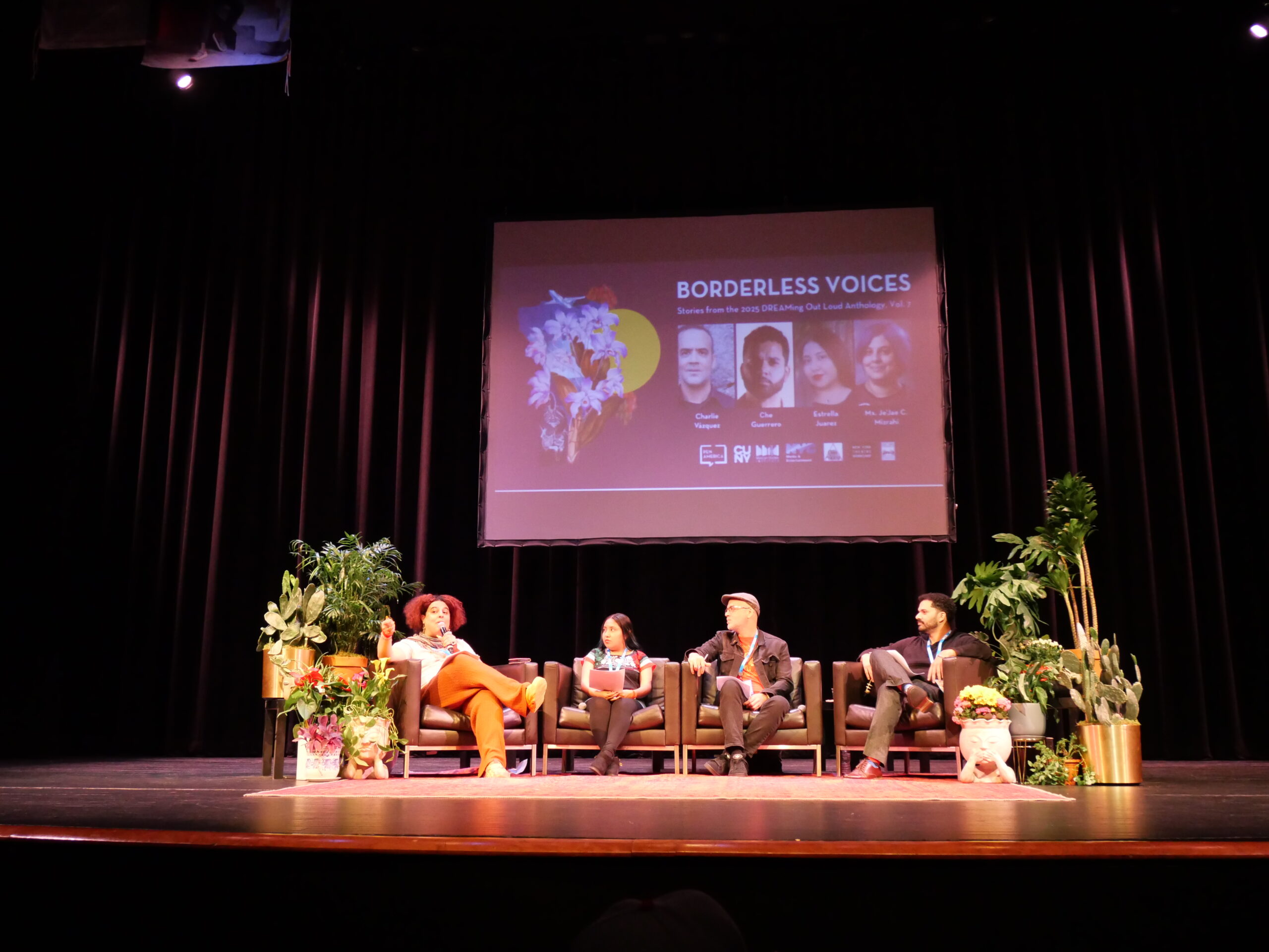 Four people sit on a stage panel discussion with plants around them. Behind them is a large screen displaying the event title Borderless Voices along with portraits and names of the panelists.