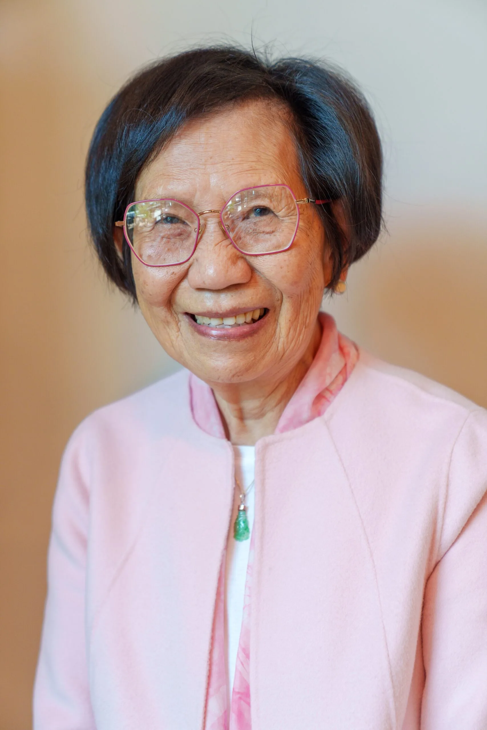 An elderly woman with short dark hair and glasses smiles warmly. She is wearing a light pink jacket over a white top, with a green pendant necklace, against a soft, neutral background.
