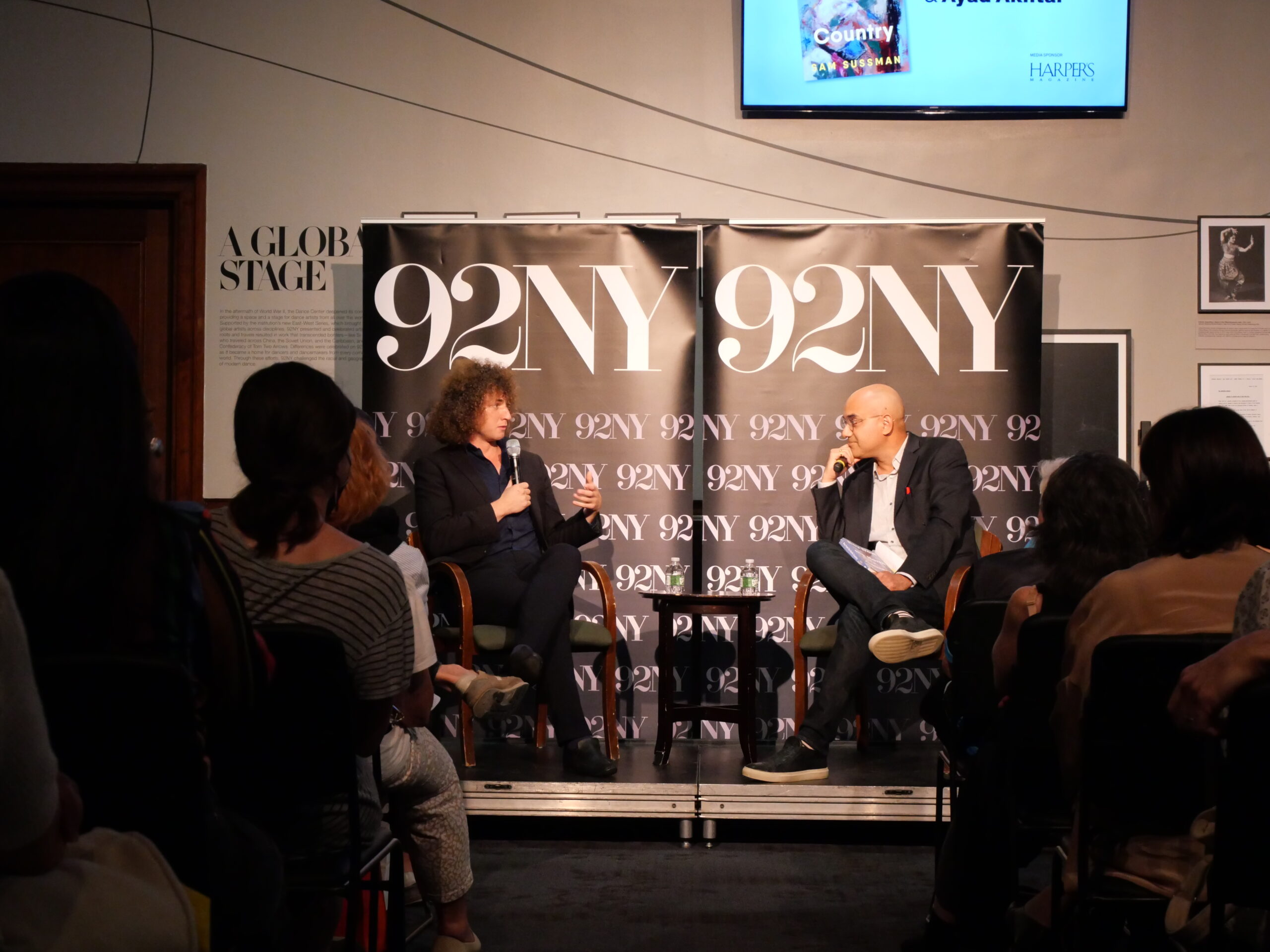Two people sit on stage in conversation at a 92NY event, facing an audience. Behind them are black banners with 92NY in white letters. The atmosphere appears to be that of a live discussion or interview.