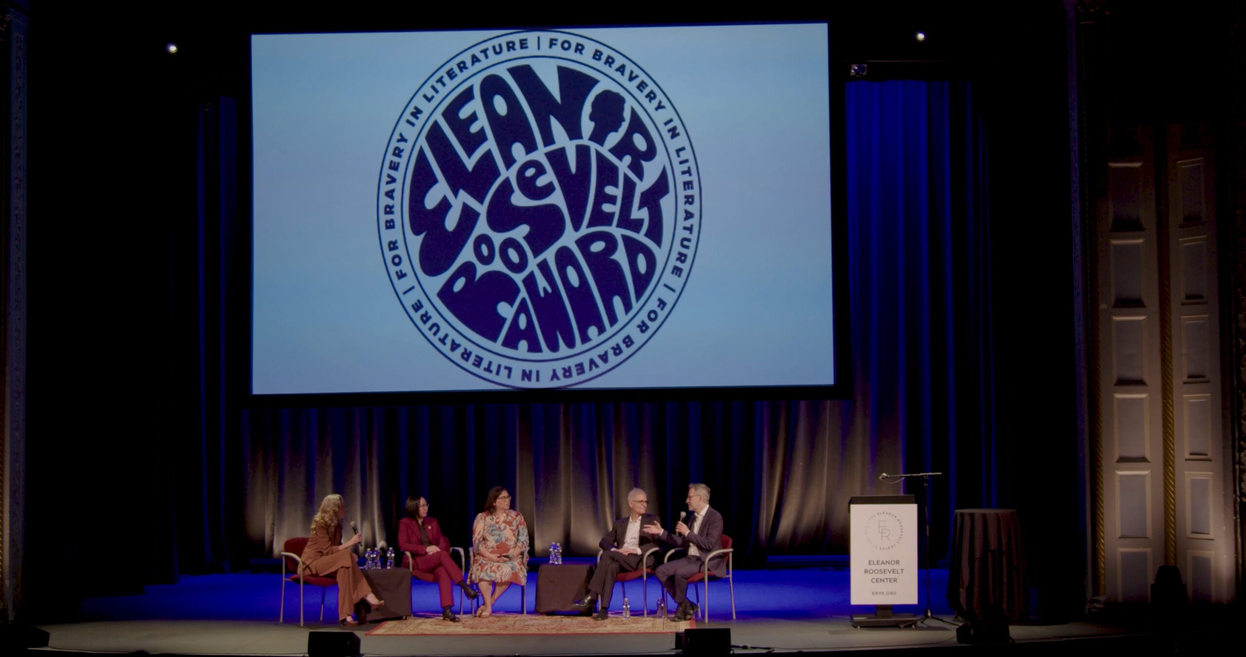 Five people are seated on stage for a panel discussion beneath a large screen displaying a circular graphic that says Lean for Braver, Level Up, Go Forward at a literature event. A podium stands at the side of the stage.