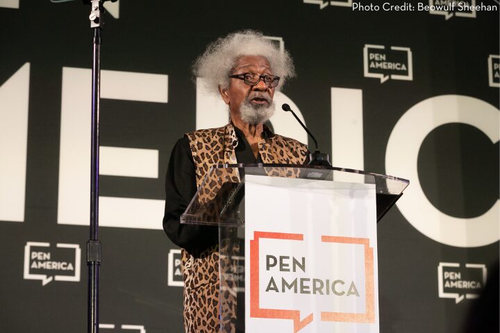 An elderly person with white hair and a beard, wearing glasses and a leopard print vest, speaks at a podium with the PEN America logo. The background features multiple PEN America signs.