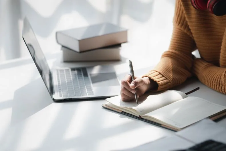 A person wearing a brown sweater writes in a notebook at a white desk with an open laptop and stacked books nearby. Sunlight casts shadows across the workspace.