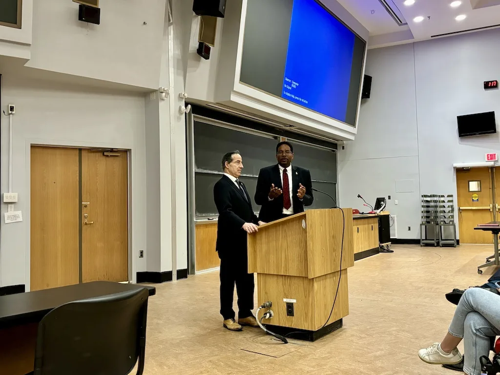 Two men in suits stand at a podium in a large lecture hall. One is speaking while the other listens. A blue projector screen is behind them, and several students are seated to the right, listening.