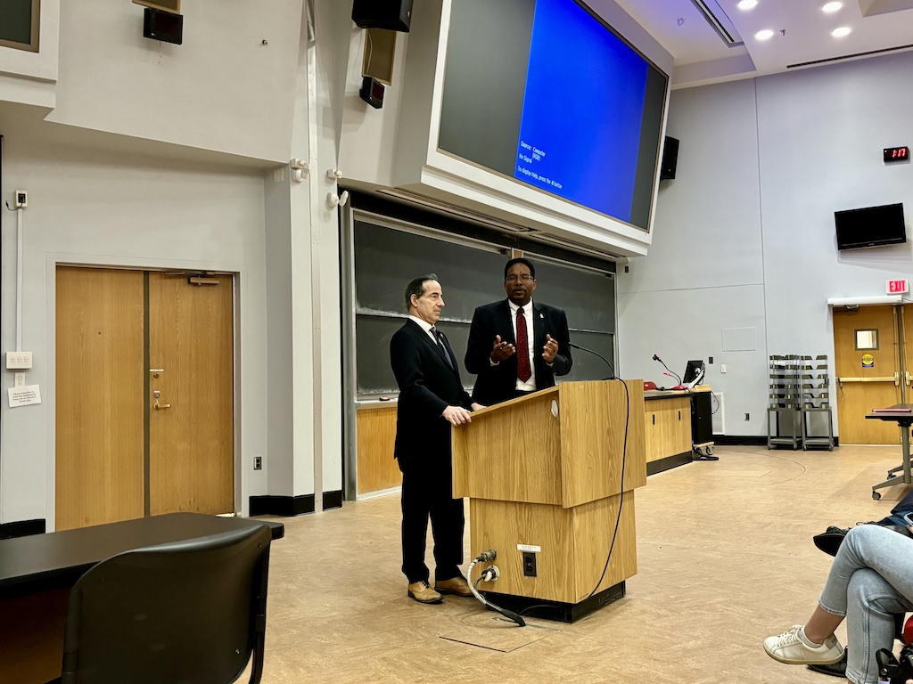 Two men in suits stand at a podium in a large lecture hall. One is speaking while the other listens. A blue projector screen is behind them, and several students are seated to the right, listening.