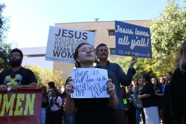 A woman holds a handwritten sign reading “There is still love in the world. LOVE THEM.” Behind her, people hold signs, one saying “JESUS VALUES AND RESPECTS TEXAS STATE WOMEN” and another, “Jesus loves all y’all!”.