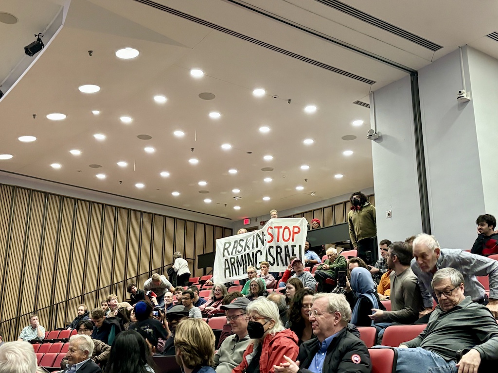 Audience seated in a lecture hall, with two people at the back holding a sign that reads Raskin STOP ARMING ISRAEL. The mood appears attentive, and some people are looking toward the sign.