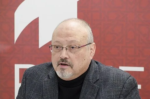 A middle-aged man with glasses, a mustache, and a goatee is wearing a black turtleneck and gray blazer, speaking in front of a red background with white geometric patterns and shapes.