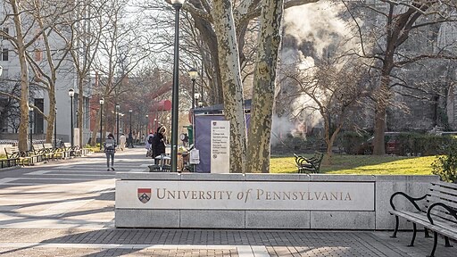 A stone sign reading University of Pennsylvania stands at the entrance to a campus walkway lined with trees, benches, and street lamps, with people walking and smoke rising in the background.