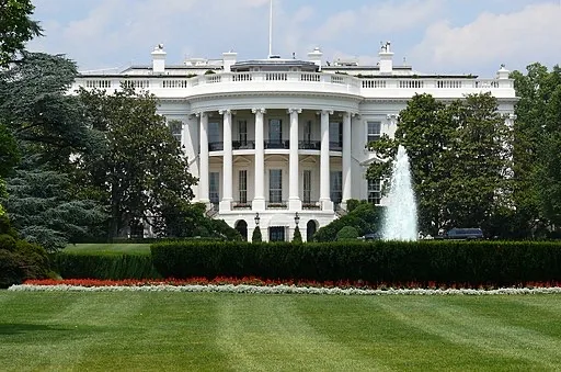 The White House, a large white neoclassical building with columns, is seen behind a green lawn, manicured bushes, a flower bed, and a water fountain spraying upward on a sunny day.