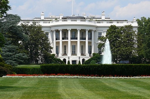 The White House, a large white neoclassical building with columns, is seen behind a green lawn, manicured bushes, a flower bed, and a water fountain spraying upward on a sunny day.